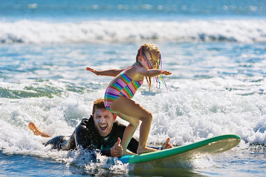 Personal Insurance - Little Surfer Girl Learning to Ride on a Surfboard With an Instructor Behind Her in the Ocean